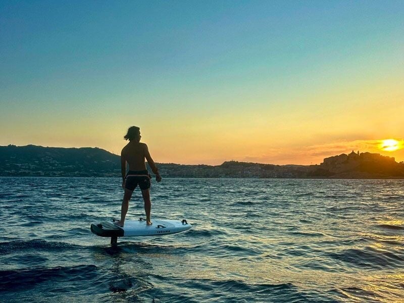 Personne debout sur une planche de wakeboard à foil au coucher du soleil avec des collines en arrière-plan.