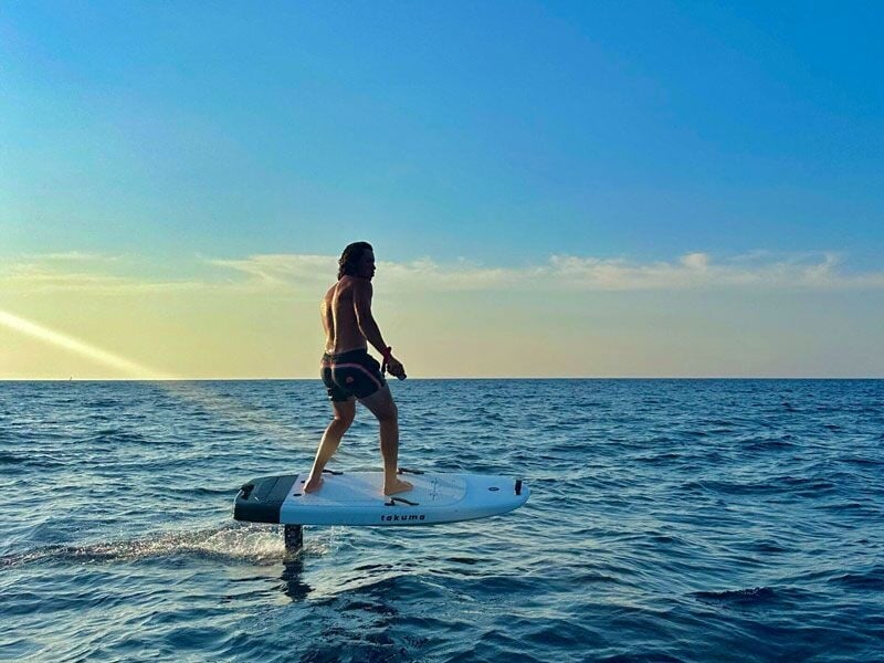 Homme en short debout sur une planche de wakeboard à foil sur une mer calme sous un ciel bleu clair.