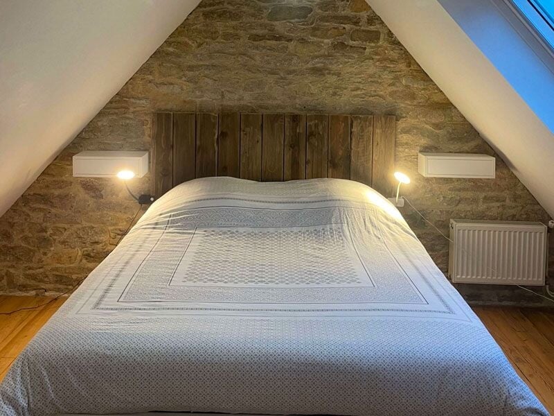 Chambre mansardée avec lit double, mur en pierre apparente et éclairage doux de chaque côté du lit.