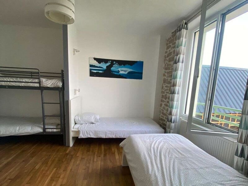 Chambre avec lits superposés et un lit simple, parquet en bois et grande fenêtre laissant entrer la lumière.