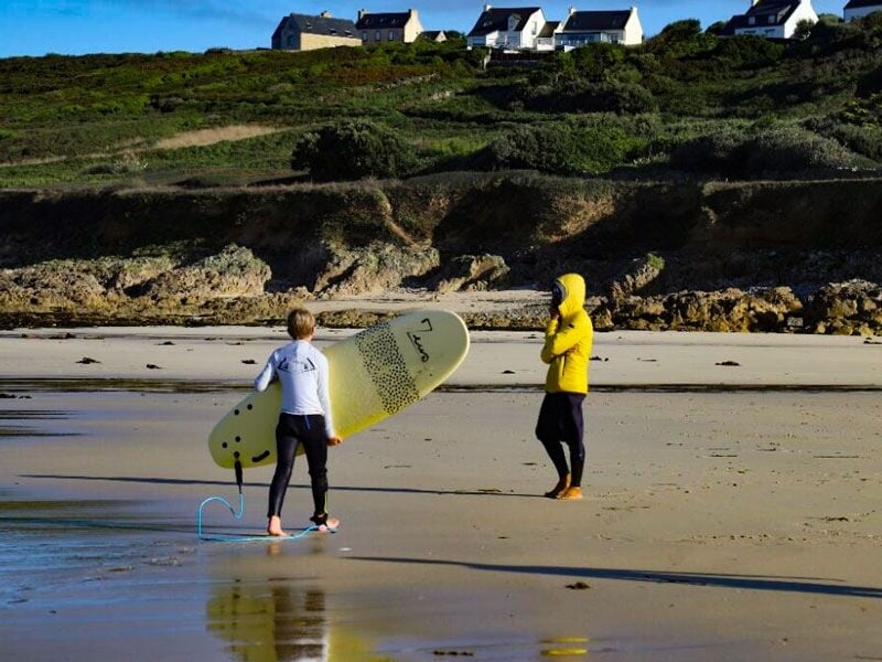 Personne en combinaison jaune discutant avec un enfant tenant une planche de surf sur une plage rocheuse.