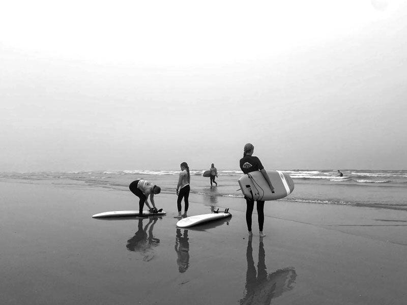 Groupe de surfeurs sur la plage, certains préparant leurs planches, ambiance en noir et blanc.