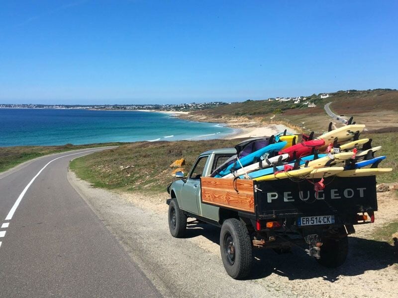 Petit camion Peugeot chargé de nombreuses planches de surf, roulant sur une route côtière ensoleillée.