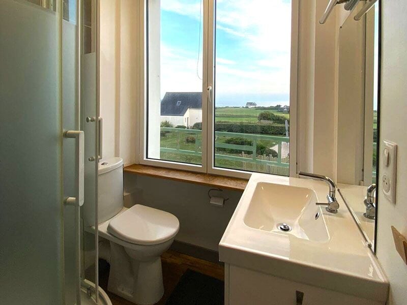 Salle de bain avec toilettes, lavabo, grande fenêtre offrant une vue sur la campagne verdoyante.