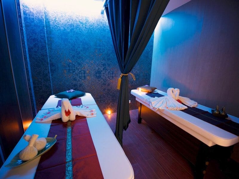 Salle de massage avec deux tables, décor bleu et rideau, ambiance zen et accessoires pour spa.