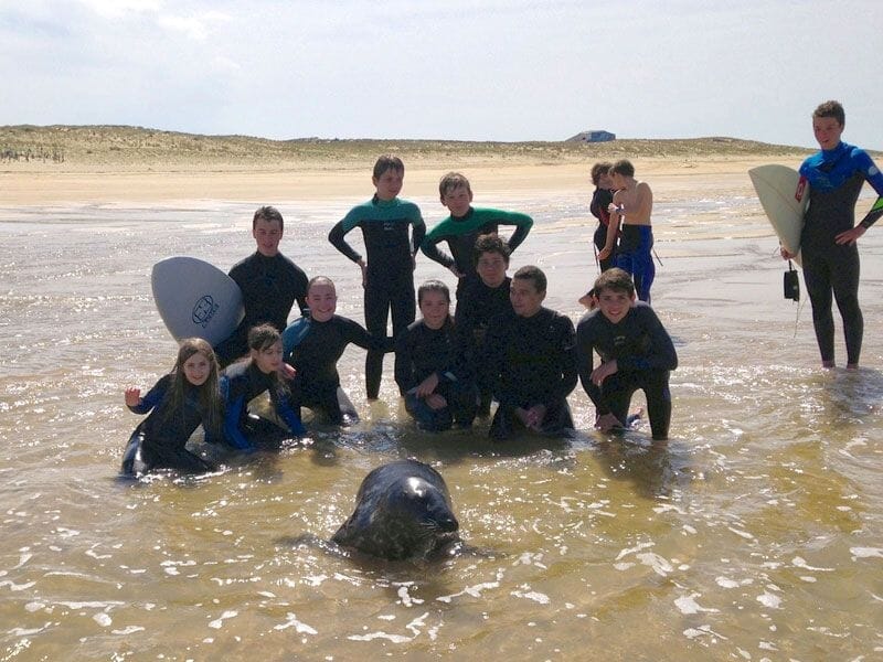 Groupe d'enfants en combinaison de surf posant dans l'eau peu profonde avec une plage et dunes en arrière-plan.