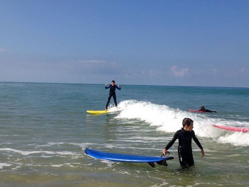 Enfants en combinaison de surf sur des planches colorées attrapant une vague calme sous un ciel clair.