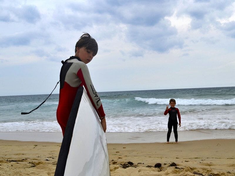 Deux enfants en combinaison de surf sur la plage, l'un tenant une planche blanche, l'autre faisant un signe de la main.