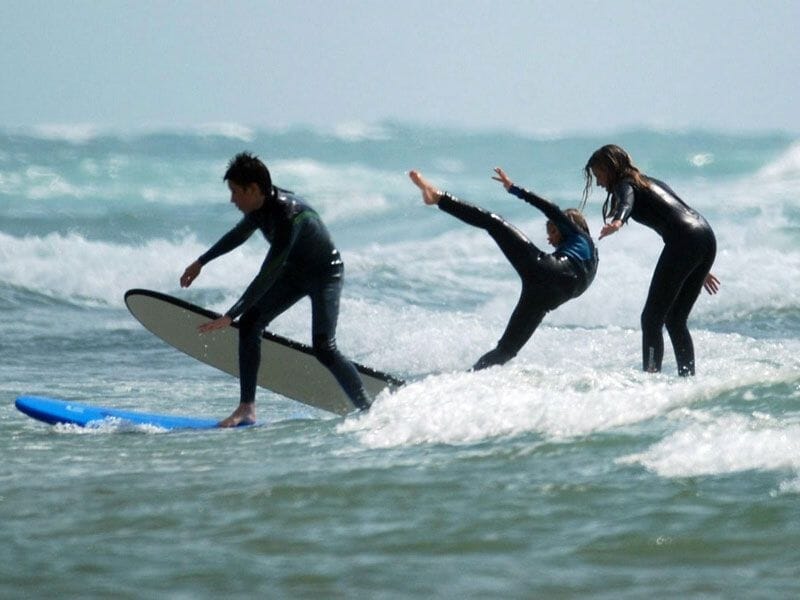 Trois enfants en combinaison de surf dans l'eau, l'un tombant d'une planche tandis que les autres essaient de garder l'équilibre.