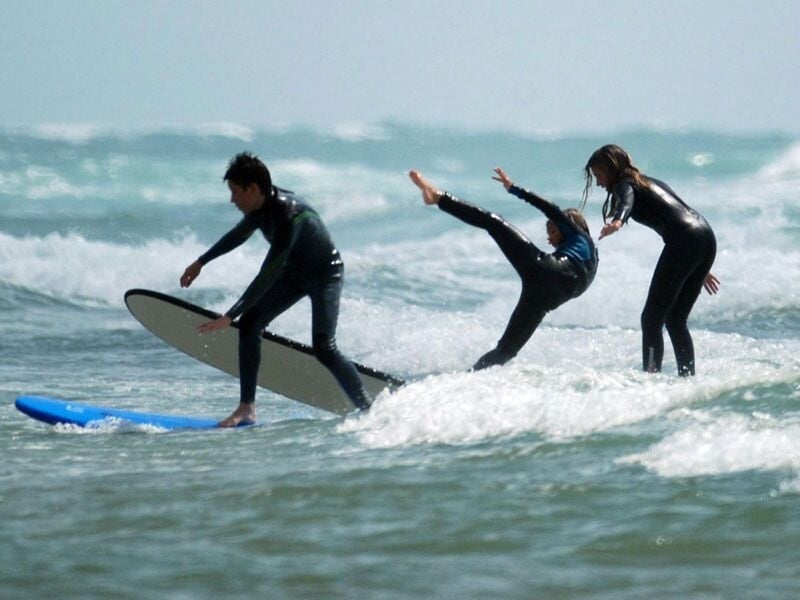 Trois enfants en combinaison de surf dans l'eau, l'un tombant d'une planche tandis que les autres essaient de garder l'équilibre.