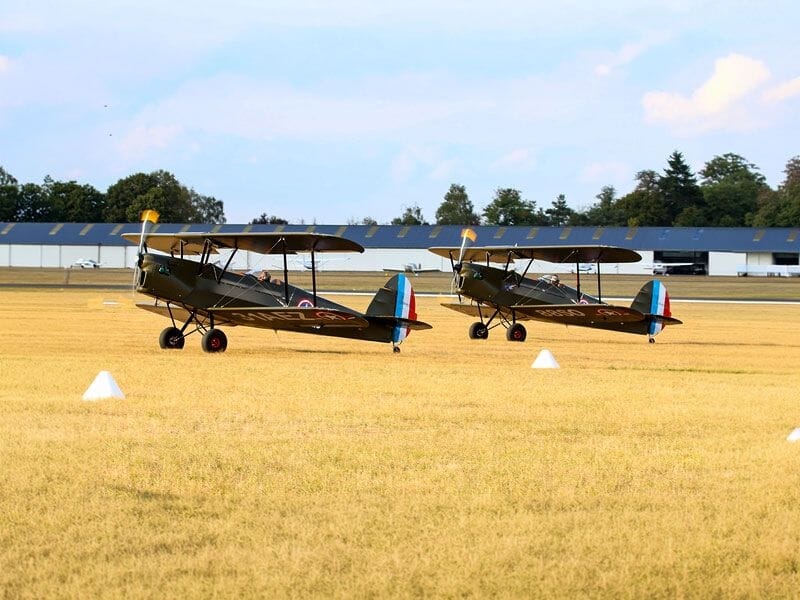 Deux avions biplans militaires anciens alignés sur un champ d'herbe jaune sous un ciel bleu clair avec quelques nuages.