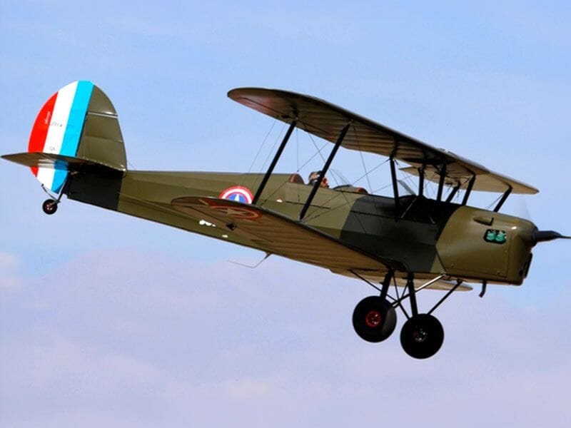 Avion biplan militaire vert en vol avec drapeau tricolore français sur la queue et ciel bleu en arrière-plan.