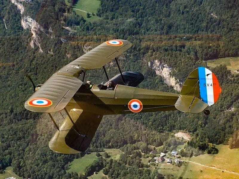 Avion militaire vintage vert volant au-dessus d'une forêt dense avec des montagnes en arrière-plan.