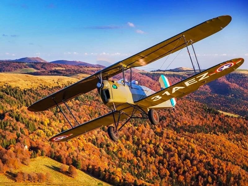 Avion biplan jaune survolant une forêt aux couleurs d'automne avec un ciel bleu clair.