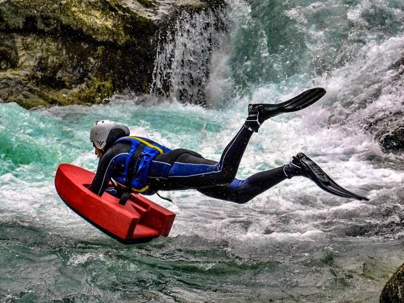 Personne en combinaison et casque blanc pratiquant l'hydrospeed dans une rivière avec eau vive et rochers environnants.