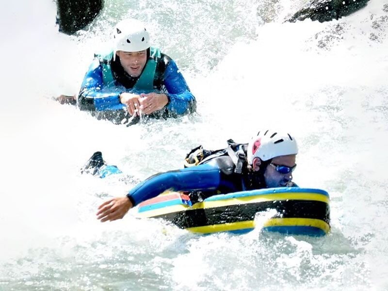 Deux personnes en casque blanc et combinaison bleue pratiquant l'hydrospeed sur une rivière agitée avec eau vive et éclaboussures.