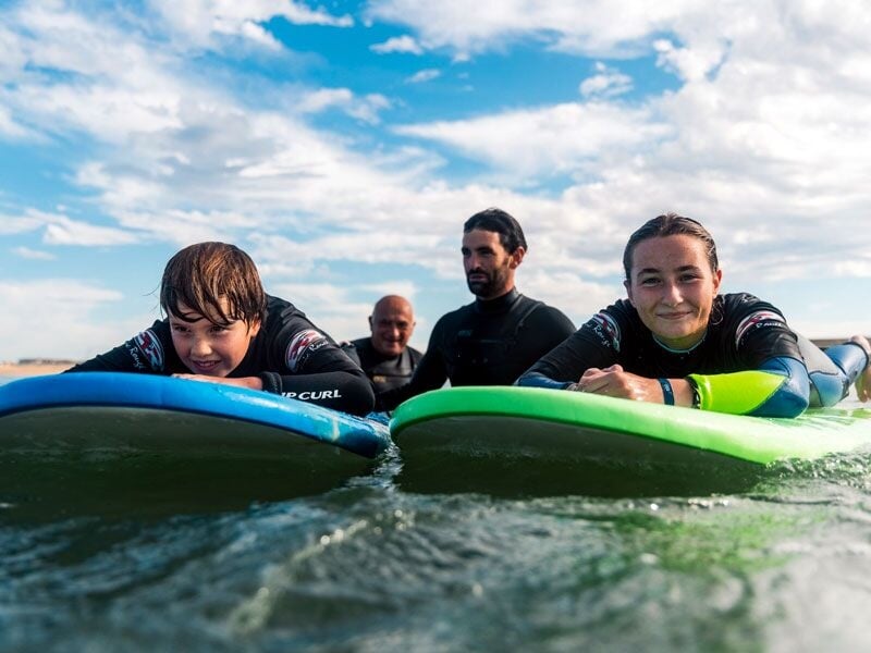 Trois surfeurs allongés sur leurs planches dans l'eau, souriants, avec un ciel bleu et nuageux.