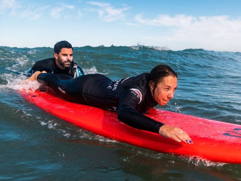 Surfeuse allongée sur une planche rouge, pagayant dans l'eau avec un instructeur derrière elle.