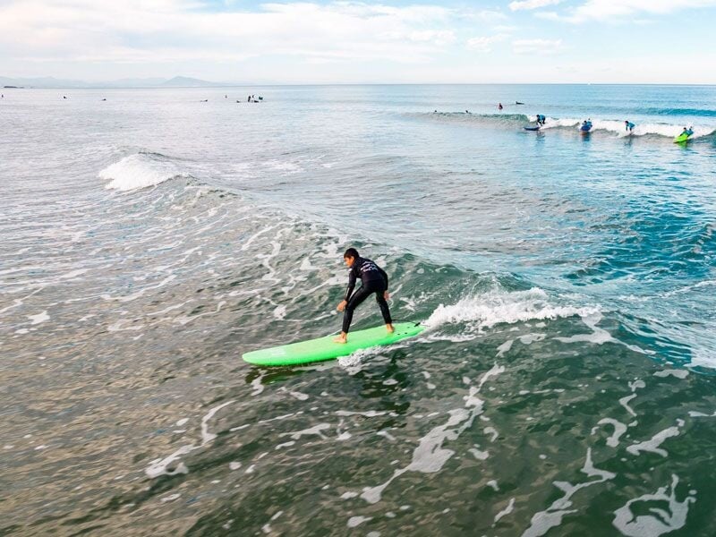 Surfeur en combinaison noire surfant sur une vague verte avec d'autres surfeurs en arrière-plan.