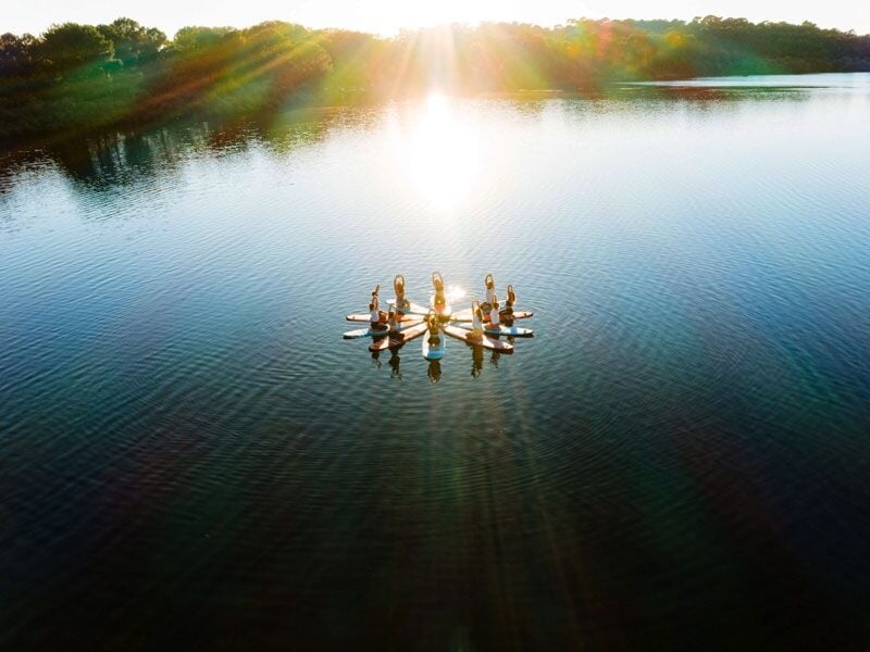 Groupe de personnes en paddle sur un lac calme au coucher du soleil, formant un cercle sur l'eau paisible.