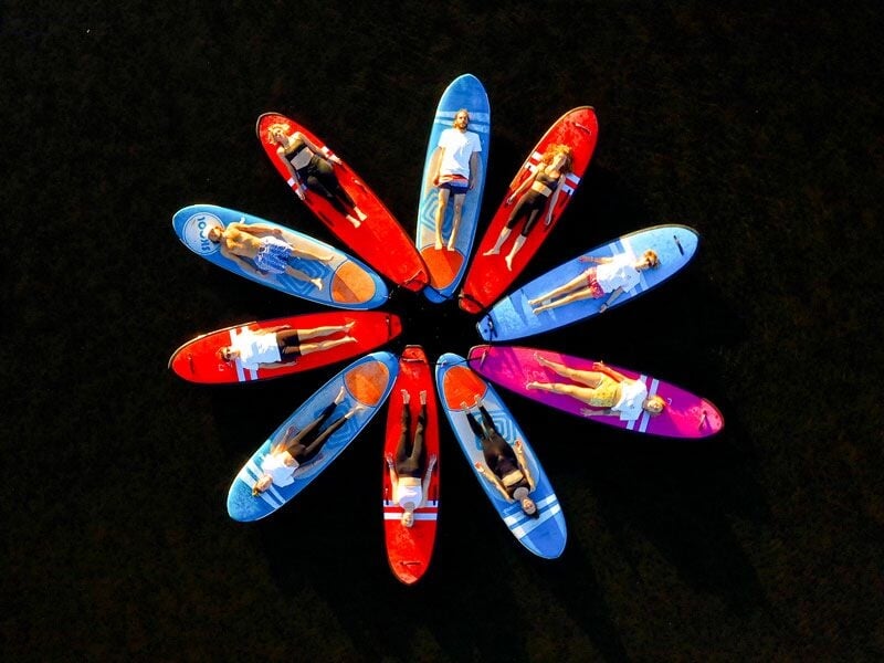 Vue aérienne de plusieurs personnes allongées sur des paddles colorés disposés en cercle sur une eau sombre.