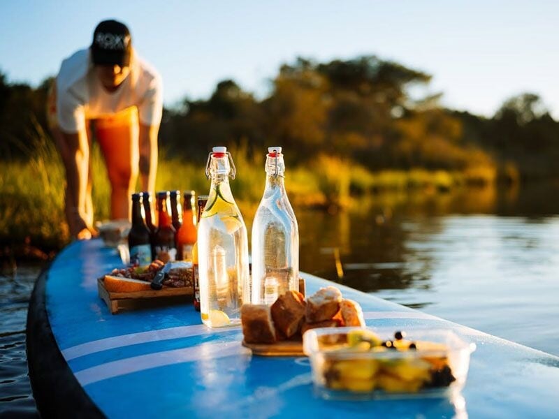 Plan rapproché sur une planche de paddle avec des bouteilles d'eau et des snacks, une personne en arrière-plan.