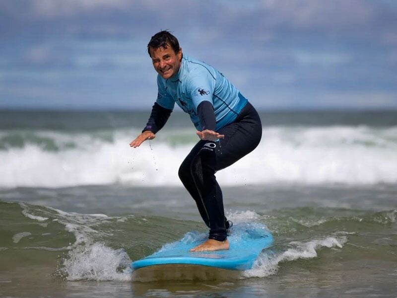 Surfeur souriant en combinaison debout sur une planche bleue, glissant sur une vague en mer.