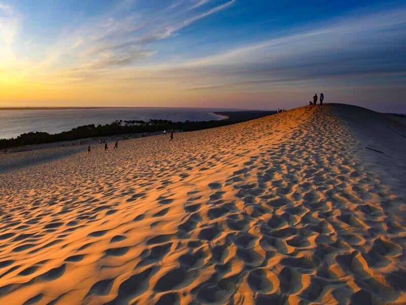 Coucher de soleil sur une grande dune de sable avec des silhouettes de personnes au sommet.