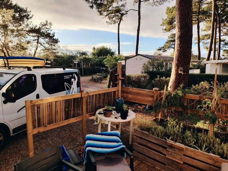 Terrasse en bois avec mobilier de jardin, plantes et un van blanc stationné à côté sous des arbres.