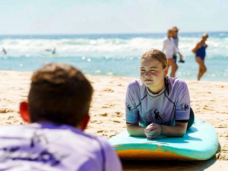 Jeune fille allongée sur une planche de surf bleue sur la plage, avec des personnes en arrière-plan au bord de l'eau.