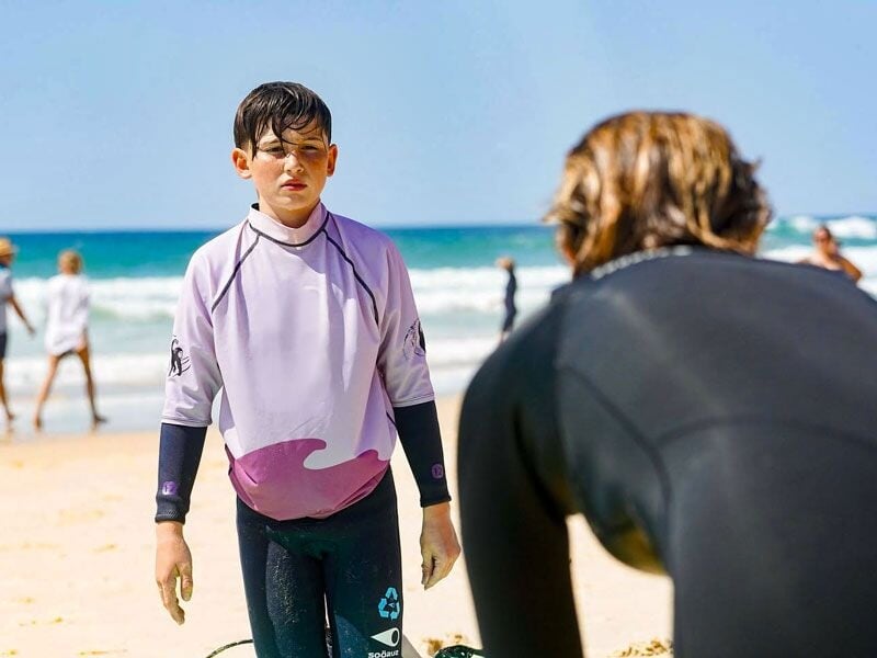Garçon en combinaison de surf debout sur le sable, regardant vers une autre personne en combinaison noire sur la plage.