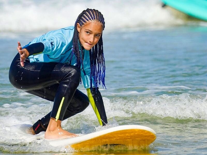 Jeune fille avec des tresses bleues en position de surf sur une planche dans l'eau peu profonde, prête à attraper une vague.