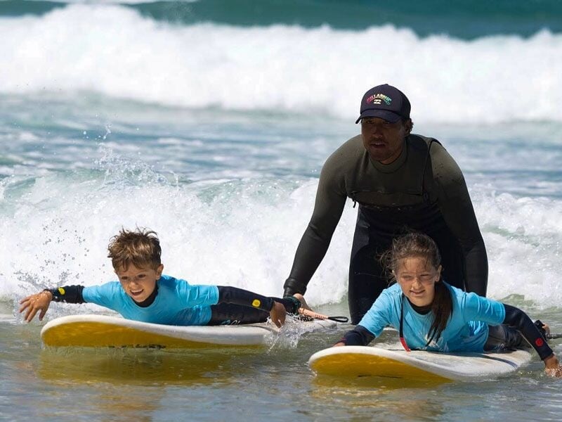 Deux enfants en combinaison bleue sur des planches de surf, aidés par un instructeur dans l'eau avec des vagues.