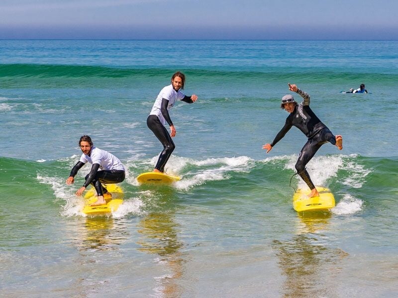 Trois surfeurs debout sur leurs planches jaunes, surfant sur une petite vague dans une mer calme sous un ciel clair.