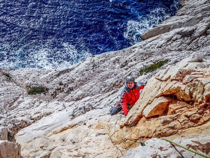 Escaladeur en veste rouge progressant sur une paroi rocheuse blanche avec la mer agitée en arrière-plan.
