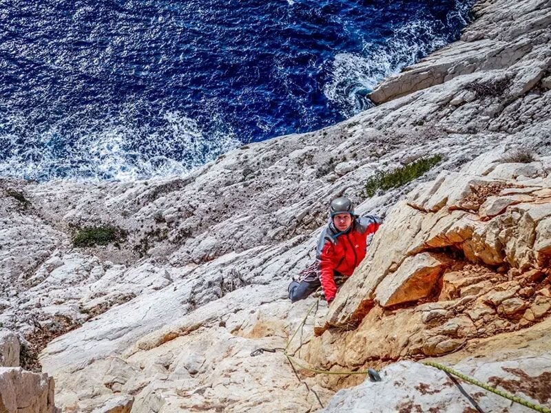 Escaladeur en veste rouge progressant sur une paroi rocheuse blanche avec la mer agitée en arrière-plan.