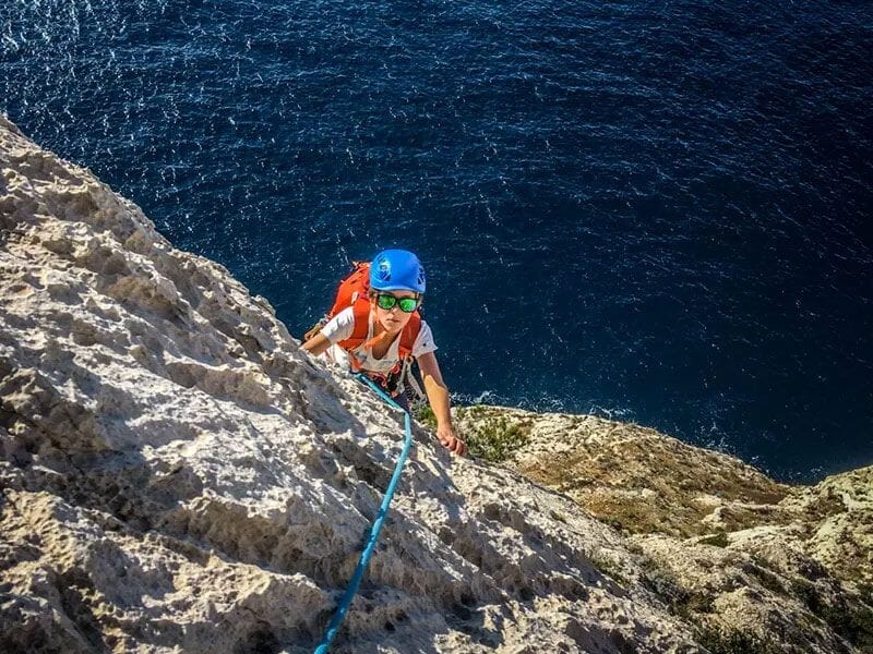 Personne en casque bleu et lunettes de soleil grimpant une falaise escarpée au bord de la mer à Marseille.