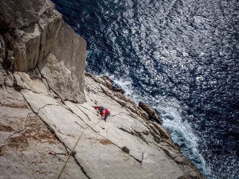 Vue aérienne d'un grimpeur en veste rouge sur une falaise blanche dominant la mer Méditerranée agitée.