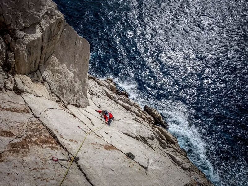 Vue aérienne d'un grimpeur en veste rouge sur une falaise blanche dominant la mer Méditerranée agitée.