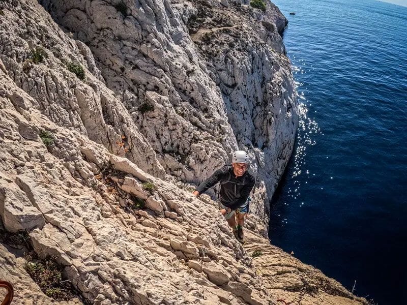 Homme en casque gris souriant en escaladant une falaise rocheuse au-dessus de la mer calme et bleue.
