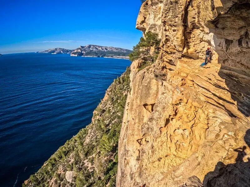 Grimpeur sur une paroi rocheuse jaune avec vue panoramique sur la mer bleue et les montagnes au loin.