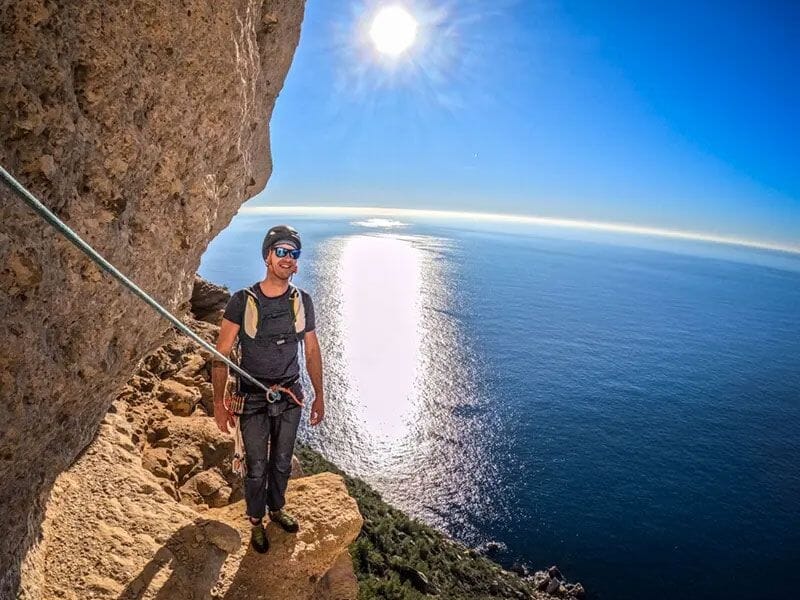 Homme en tenue noire et casque souriant sur une corniche rocheuse avec le soleil brillant sur la mer.