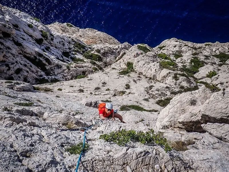 Escaladeur en veste rouge grimpant une falaise blanche escarpée avec la mer Méditerranée en contrebas.