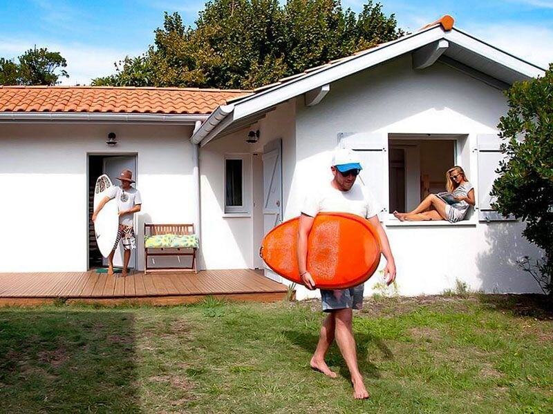 Un homme marche sur l'herbe avec une planche de surf orange, une femme est assise à la fenêtre d'une maison blanche.