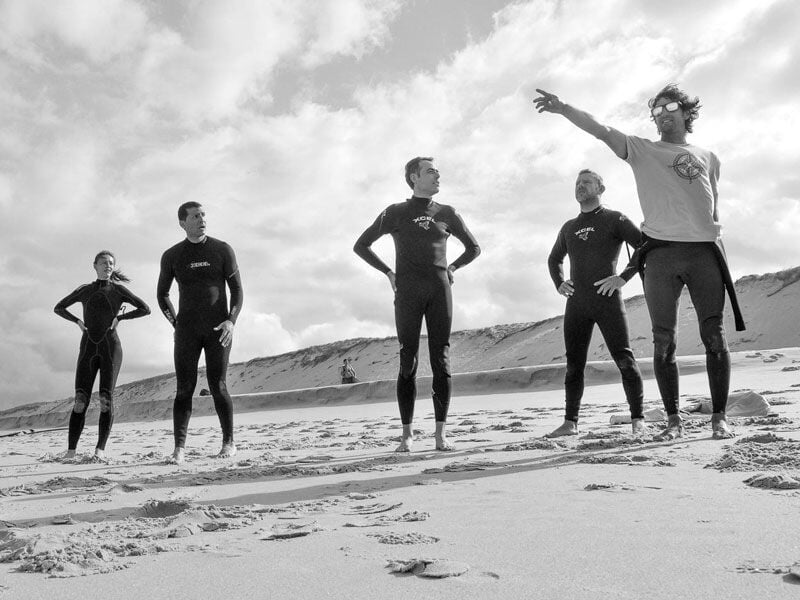 Cinq surfeurs en combinaison noire posent sur la plage, un d'eux pointe du doigt vers l'horizon, photo en noir et blanc.