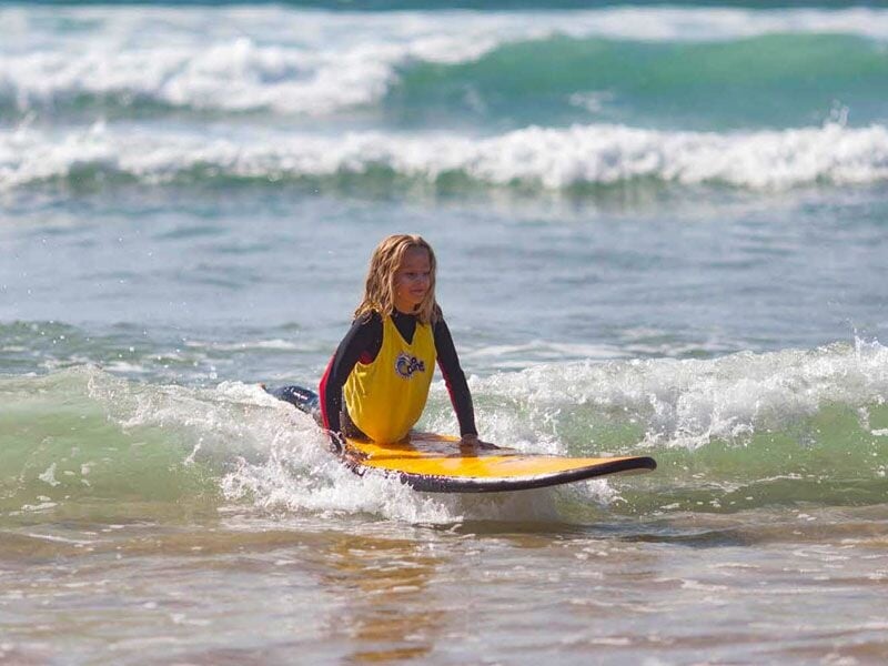 Un enfant en combinaison jaune et noire apprend à surfer sur une petite vague avec une planche jaune dans l'eau.