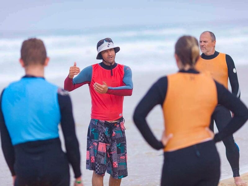 Un instructeur en combinaison rouge parle à un groupe de surfeurs en combinaison noire et orange sur la plage.