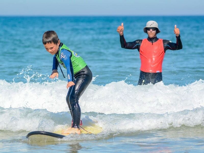 Un enfant en combinaison bleue surfe sur une petite vague, un adulte en combinaison rouge fait un pouce levé dans l'eau.