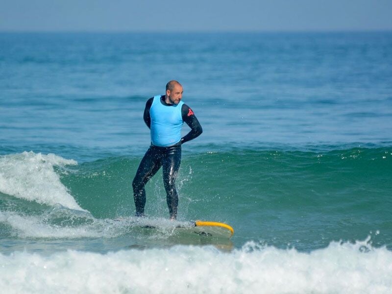 Un homme en combinaison bleue surfe sur une vague de taille moyenne dans une mer calme sous un ciel clair.