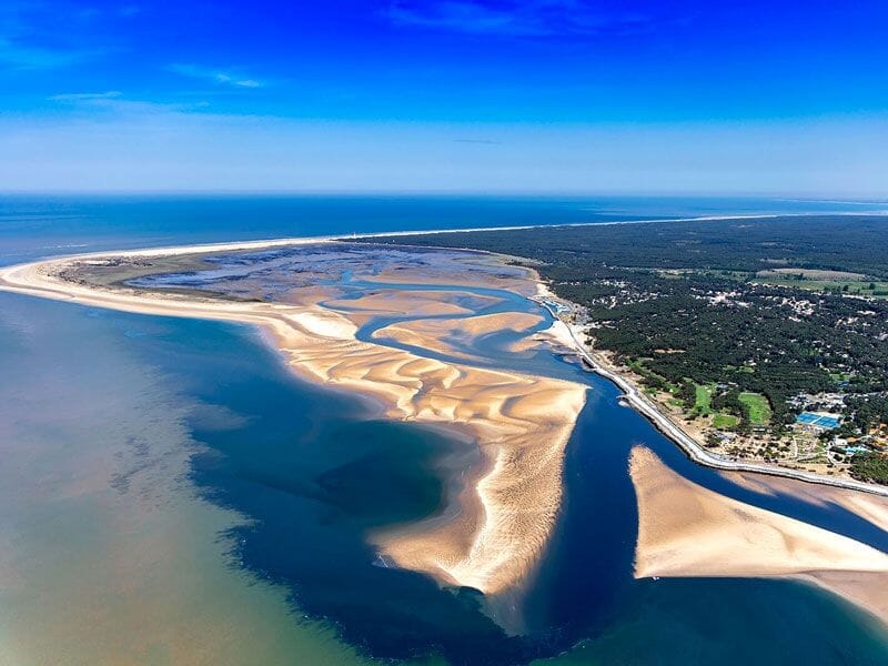 Vue aérienne d'un estuaire avec bancs de sable et eau turquoise sous un ciel bleu clair en bord de mer.